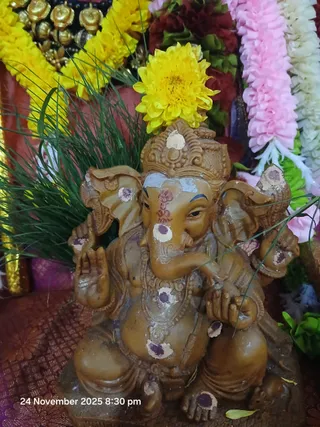 Close-up photo of a brown clay murti of Lord Ganesha seated on a pedestal, adorned with yellow chrysanthemum flowers on its head, green grass blades, and white and purple dots of vibhuti/kumkum on its trunk, forehead and limbs. Behind it are garlands of yellow marigold and pink and white jasmine strands, and a red fabric draped beneath. A timestamp in the corner reads 24 November 2025 8:30 pm. No people are visible.