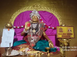 A golden statue of a seated figure is the central subject, set before a gold arched frame and magenta drapery. The figure wears a white and gold turban, a red bindi, layered rudraksha and bead necklaces, a large pendant, and a brown floral garland, with a teal and maroon silk garment. The right hand is raised in blessing holding a red flower. Brass oil lamps, a kalasha, a bowl of white petals and incense sticks sit on the altar. A yellow sign reads KAILASA SALEM BHARATH beside a small portrait. The statue is a murti, not a living person.