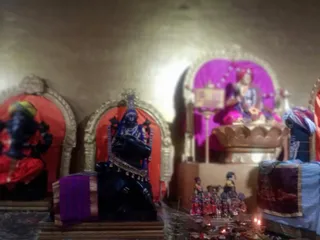 Blurred photograph of a shrine room with several deity murtis in gilded arched niches lined with red and magenta cloth. On the left, a dark murti wears red and green garments. In the centre, a black stone murti sits on a bull-shaped base draped with purple cloth and gold trim. On the right, a figure in a magenta-lit niche sits on a golden peetha behind a small framed placard. Small clay oil lamps burn on the floor beside a row of tiny painted figurines in red and blue, and a blue and gold cloth hangs at right. No people are visible.