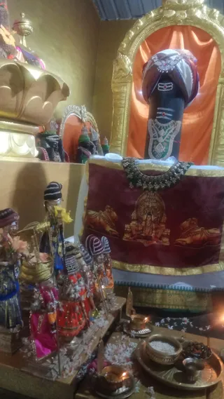 A photograph of a temple shrine interior. Centre right, a tall black stone lingam wrapped in a patterned turban-like cloth with a red gem and white tripundra lines is set in a gold arched niche against an orange satin backdrop. It wears a silver and green-beaded necklace and sits before a maroon embroidered cloth showing a seated deity with bulls. On the left, several doll-like murtis in red, pink, and blue dresses, with striped headdresses, stand on a ledge scattered with white flowers. In the foreground, brass oil lamps, a brass plate with rice, and a small cup sit on a table with petals, and a lit flame glows at the lower right. No people are visible.