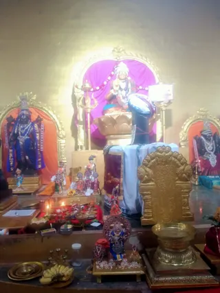 Photograph of a temple shrine with no people visible. A central seated deity murti with a white turban and garland sits on a golden lotus base before a magenta backdrop, beside a tall trident. A tall dark blue shivalinga-style pillar draped in white and maroon cloth stands in front. Flanking shrines hold a dark blue standing deity on the left and a dark deity with silver necklaces on the right. A brass pot, brass plates, bananas, small oil lamps with flames, and offerings in bowls sit on a red cloth in the foreground.