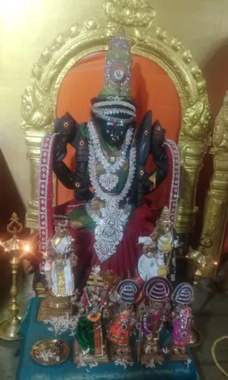A photograph of a dark-stone deity murti seated on a red cushioned throne-like seat in front of an orange backdrop and a gold arched frame. The black-faced deity wears a tall jewelled crown, layered silver and white garlands, a green and red garment, and has multiple arms. Smaller figures in white and gold attire and seated smaller murtis with purple and pink garments stand at the base, with white flower petals scattered on a turquoise cloth. A brass oil lamp burns to the left and a brass plate holding offerings sits in the foreground. No physical people are visible.