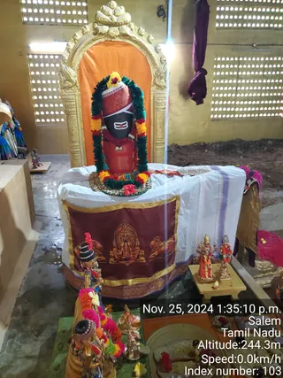 A photograph inside a temple shrine showing a Shiva lingam with a reddish-orange cylindrical base and a black stone top marked with white tripundra lines and a red dot, adorned with green and orange-yellow marigold garlands and a red flower. The lingam stands within a gilded arched niche with an orange backdrop, on a white cloth draped over a platform. A maroon cloth with gold border at the front shows a painted seated deity flanked by bulls. Smaller brass figures and a brass oil lamp with flower offerings sit at the front, and a pair of small deity figures stand on a wooden stand to the right. No people are visible. Timestamp overlay reads Nov. 25, 2024 1:35:10 p.m., Salem, Tamil Nadu.
