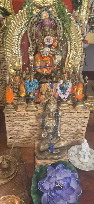 A photograph of a home shrine with a golden arched frame draped in green vine garlands. At its centre stands a brass deity murti in an orange and gold cloth, adorned with a multicoloured flower garland and a jewelled peacock-feather crown ornament. Smaller brass figures in orange cloth flank it on a woven wicker platform. In front sits a brass seated deity murti with a rudraksha mala and a trident-like staff, beside a blue lotus-leaf flower decoration, a brass oil lamp with lit incense, and a white figurine on a patterned plate. A small printed picture of several deities is tucked behind the smaller figures.