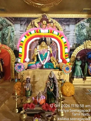A golden deity murti of Meenakshi seated on a raised golden pedestal beneath an arch of red, yellow, white and green flower garlands with pink edging. The goddess wears a teal and lavender saree, a black crown and layered gold necklaces, and her raised right hand shows a blessing gesture. Smaller golden murtis stand in front, including one in green and gold clothing and a green-robed figure holding a staff. Brass oil lamps, a kalasha and red hibiscus flowers surround the altar, and a painted temple backdrop with trees and stone carvings fills the background. A timestamp in the corner reads 12 Oct 2026 11:19:58 am, Periya Seeragapadi, Tamil Nadu, kailasa seeragapadi.