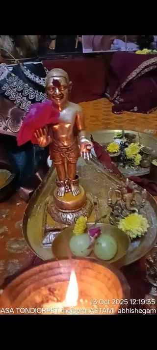 A photograph of a small gold-coloured metal standing figure of a child-like deity murti with a red tilak mark on the forehead, one hand raised in blessing, placed on a brass tray with a yellow marigold garland, a small Ganesha figure, lemons or limes, a pink flower and a lit oil lamp in the foreground. Behind it are maroon and embroidered cloth draped over a table, and a framed picture at the top. A timestamp reads 13 Oct 2025 12:19:35 with the caption KAILASA TONDIORPET theiperai ASTAMI abhishegam. No people are visible.
