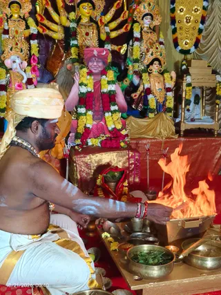 A kneeling priest in a cream-coloured turban and white dhoti with a gold border performs a homa at a fire pit in front of a decorated shrine. He is bare-chested with white ash markings on his arms and wears a beaded necklace and red-and-white bangles. Flames rise from a metal fire box on a brass-edged wooden tray, with brass bowls of green leaves, petals and small oil lamps arranged around it. Behind him stands a shrine with yellow and white marigold garlands, multi-armed deity murti including a Ganesha figure, and a seated figure in a red turban with a garland and red-and-yellow flower garlands, its face painted. The figure is an image rather than a living person, so the scene has one physical person.