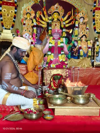 A priest with a cream turban, bare chest, white dhoti with gold border and white sacred markings on his arms and chest sits on a red cloth beside a brass homa setup. A flame burns in a copper fire box on the right, with brass bowls, a kalasha, marigold and rose offerings, and small oil lamps on the floor. Behind them, a large framed image of a seated figure in a red-and-pink turban and red garment, adorned with a garland of yellow, white and red flowers, has raised hands. Flanking this are colourful multi-armed goddess murti and a figure with a golden crown and garlands, all decorated with flower garlands. The scene is a Navaratri ritual with a homa at KAILASA Hyderabad.