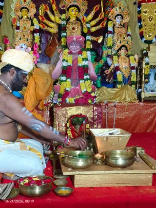 A kneeling priest with a bare chest, white sacred ash markings on his arms and chest, a cream turban and a white dhoti with gold border performs a ritual before a decorated shrine. He tends brass bowls holding green leaves and a small oil lamp on a wooden platform covered in red cloth, with a wooden box beside them. Behind him stands a large framed printed portrait of a smiling figure in a red-orange turban and red robes, its hands raised in blessing, garlanded with yellow, white and red flower strands. Around it are multi-armed deity murtis with gold crowns and flower garlands, and a brass bowl of rose petals sits in the foreground. A second person in orange robes is partly visible at left. The portrait is a printed depiction, not a physical person, and a timestamp reads 10/21/2026, 00:19.