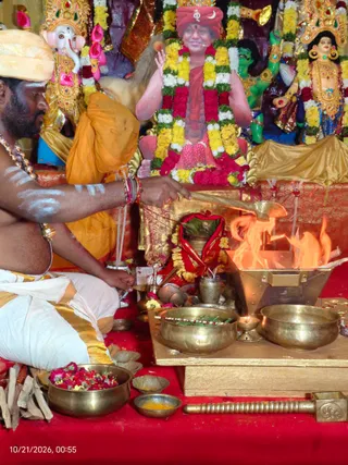 A priest seated on a red cloth conducts a homa at a metal fire pit, pouring an offering with a long brass ladle into flames. He wears a white dhoti with gold border, an orange shawl, a gold cloth headwrap and a beaded necklace, with white sacred markings on his bare arms and chest. Brass bowls hold flower petals, leaves and offerings, and a brass pestle lies in front. Behind him, a large framed portrait of a smiling figure in a red and orange robe and red turban with a crescent emblem, garlanded with yellow and red flowers, stands among colourful deity murtis of Ganesha and a goddess with gold crowns. The Hindu homa ritual is the main subject.
