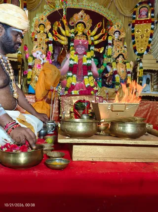A photograph of a Navaratri worship scene in a temple-like setting with gold drapes and red cloth. On the left, a bare-chested priest in a white dhoti with a cream turban and beaded mala necklaces holds a brass bowl of red flowers and sits before the altar. Behind a brass fire vessel with a bright flame, a central figure in a red turban and red garments sits before a multi-armed goddess murti adorned with yellow and red garlands, flanked by Ganesha and other deity murtis. A framed portrait with a saffron turban hangs at upper right, along with a garland-decorated picture. Brass bowls, a lamp, leaves and an offering pot sit on the red-covered platform. Visible people: the priest and the seated figure in red.