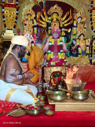A photograph of a homa ritual at a temple-like shrine. A priest with a white and gold turban, bare-chested with white sacred markings and a beaded necklace, sits cross-legged in white dhoti on a red cloth, tending a fire in a brass fire pit at right. Brass bowls with leaves and offerings, a small oil lamp and flower garlands sit on the red floor. Behind him stands a large framed portrait of a seated figure in red and pink garments with a red turban and marigold garlands, flanked by multi-armed yellow deity murtis in ornate crowns and gold jewellery, with a ganesha figure to the left. A gold-embroidered banner hangs above. Two people are visible: the priest and a standing assistant in orange cloth.