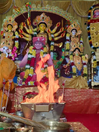A seated priest in a red-and-orange turban with a crescent emblem and a red shawl performs a fire ritual before a multi-armed deity murti with a crown and gold-trimmed decor, flanked by other painted deity figures, garlanded with marigold and red flower garlands. In the foreground a flame rises from a brass homa vessel, with brass ladles and bowls around it. A framed portrait with a garland hangs at the right edge. A timestamp in the corner reads 10/21/2026, 01:20.