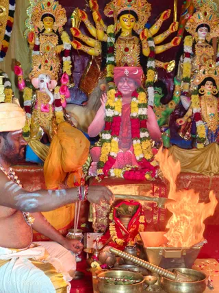 A homa ritual at a shrine decorated with large painted deity murtis of multi-armed goddesses wearing gold crowns and jewelled ornaments, with a Ganesha figure at left. A bald priest with white-painted arms and chest and a saffron cloth over his shoulder sits at left, pouring offerings into a fire from a brass vessel; a copper ladle lies on the floor and brass bowls sit in the foreground. Central to the frame is a portrait-sized printed image of a seated figure in red-pink robes wearing a red turban with a crescent emblem, adorned with a garland of red, yellow and white flowers, raising one hand in greeting. The flames of the fire pit rise at right beside a red-draped kalasha with a garland.