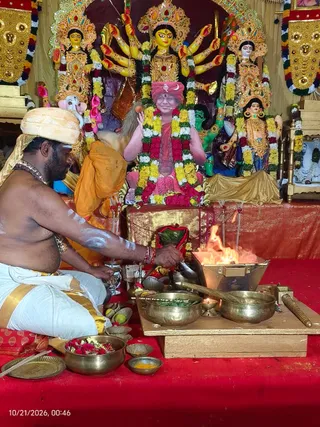 A shirtless priest with white ash markings on his arms and chest sits on a red cloth before a fire pit, pouring offerings into a flame from a brass vessel. He wears a white dhoti with a gold border and a saffron-orange cloth draped over his head. Behind him stands a multi-armed goddess murti in gold and red ornaments, flanked by additional deity figures, including a Ganesha figure with a kumkum-marked trunk, all garlanded with marigolds and roses. Brass bowls of flower petals, kumkum, leaves and small oil lamps surround the homa setup. A large framed image in the center shows a figure in red garments with a red turban bearing a crescent emblem, seated and garlanded, with its hands raised, and is a printed or painted depiction rather than a physical person. Yellow and green decorative hangings frame the altar.