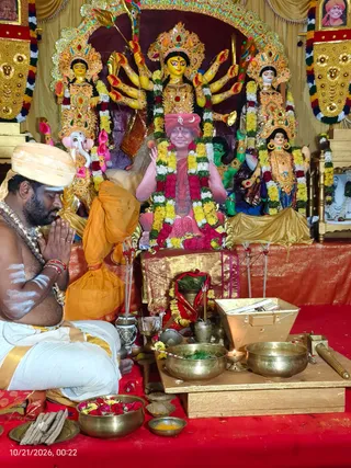 Photograph of a temple shrine during Navaratri worship. In the foreground a bare-chested priest with white body markings and a cream turban sits with palms pressed together in namaste, wearing a white dhoti with gold border and a beaded necklace. Behind him a golden-orange cloth bundle rests on the floor. The central backdrop is a multi-armed goddess murti with a gold crown holding weapons, flanked by two other painted deity figures, a Ganesha figure and a green figure, all garlanded with yellow, red and white flowers. In front of the central murti a framed image of a smiling figure in a red turban and saffron robe, wearing a rudraksha-style mala, is adorned with a large flower garland and placed on a golden-bordered red cloth. Brass bowls with flower petals, a kalasha-like vessel, lit oil lamp and brass puja implements sit on a red cloth on a wooden platform. Gold-framed portraits appear at the top corners.