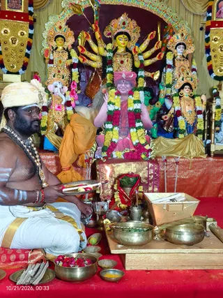 A priest seated on a red cloth at a puja altar, wearing a cream-and-gold turban, white dhoti with gold border, bare upper body, rudraksha-style beaded necklaces and white and red markings on his forehead and arms. He holds a brass plate with a lit oil lamp and flowers, while brass bowls, a flower-filled vessel, a wooden box and small offering dishes sit before him. Behind him stands a large multi-armed goddess murti in yellow and gold with a crowned head, flanked by smaller painted deity figures including an elephant-headed form, decorated with garlands in red, yellow and white. Central to the backdrop is a seated figure in a red turban and saffron robe garlanded in yellow and red blossoms, with hands raised in greeting, presented as a portrait or murti on a gold-draped throne. Gold and red drapes frame the scene.