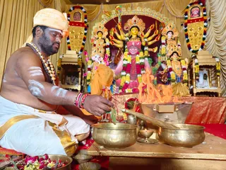 A bare-chested priest in a white dhoti with a gold border sits at a low wooden table performing a homa, placing offerings into a flame from a fire pit. He wears a rudraksha mala and white markings on his arms and forehead, with a saffron cloth over his shoulder. Behind him stands a richly decorated deity murti with multiple arms, red and gold garments, and garlands, flanked by other deity figures and framed by golden drapery. Brass bowls, a ladle and flower offerings are on the table, and a small oil lamp sits nearby. The timestamp 10/21/2026 appears in the lower left corner.