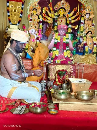 A seated priest with a bare chest, white sacred ash stripes, a cream turban and a beaded necklace performs a puja before a decorated shrine. He sits on a red cloth with his hands folded, facing brass bowls of flowers, leaves and a small lamp. Behind him stand gold-and-yellow painted deity murti: a multi-armed goddess with a crown, a Ganesha figure, and other figures, all garlanded with yellow, white and red flowers. A pink and red framed image of a seated figure in a red turban and saffron garments, garlanded with flower chains, sits in the centre of the altar, with a small golden sign on the turban. Incense sticks and a brass urn sit in front. The timestamp reads 10/21/2026.
