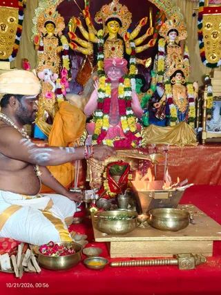 A shirtless priest with white sacred markings on his arms and chest, wearing a cream turban, white dhoti with gold border, and a gold necklace, sits on red cloth performing a fire ritual. He holds a ladle over a metal fire pit with visible flames, beside brass bowls, a flower-filled brass dish, and incense sticks. Behind him stands a framed portrait of a smiling figure in red and saffron robes with a red turban, garlanded with yellow and red flowers, flanked by colourful multi-armed goddess statues adorned with gold jewellery and garlands. A gold-trimmed drape hangs above. Scene suggests a homa in a temple setting.