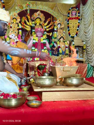 A photograph of a homa ritual at a decorated altar with a central goddess murti having multiple arms and holding weapons, flanked by Ganesha and another deity figure, all draped with yellow, white and red flower garlands. In the foreground a priest with white body markings and a white turban-style cloth tends a fire rising from a metal fire vessel, with brass bowls, rose petals and leaves in front. Behind the murtis a framed portrait with a red-and-white garland hangs on gold curtains. A seated figure in red-orange garments sits before the deities, and a visible person in a white dhoti is at left. The timestamp in the corner reads 10/21/2026, 00:38.