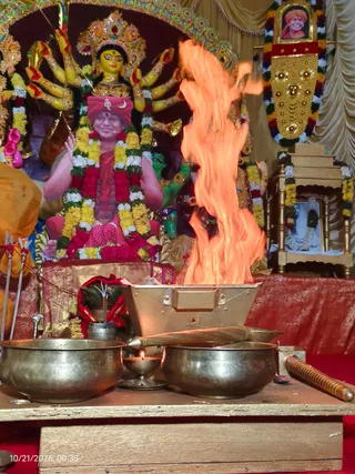 A brass homa fire pit with a tall orange flame rising from it sits in the foreground on a wooden table, flanked by two brass bowls, a brass ladle, a small oil lamp and a small kalasha-like pot wrapped in red cloth. Behind the fire, a large multi-armed deity murti with a golden crown and yellow face is adorned with marigold and flower garlands. In the background, a large printed portrait shows a smiling figure in a red-and-saffron turban with a crescent emblem, wearing a red shawl and a garland, seated on a red-cushioned throne-like seat; the portrait is printed, not a physical person. A framed photo on a small wooden shrine at right is also garlanded. No physical people are visible.