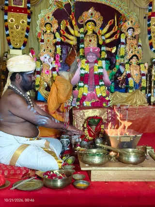 A shirtless priest in a white dhoti with a gold border and a cream turban sits on a red cloth before a homa fire, pouring offerings from a brass vessel. Behind him, a multi-armed goddess murti in gold and red crowned attire stands beside Ganesha and other deity murtis, all garlanded with marigolds and roses. Framed by gold and tassel-hung drapes, a central seated figure in a red turban and pink-and-red garlands, shown in a painted or printed form, is raised before the murtis. Brass bowls, flower offerings, a small oil lamp and a lit fire pit with flames sit on the red-draped platform. A date stamp in the corner reads 10/21/2026, 00:46.