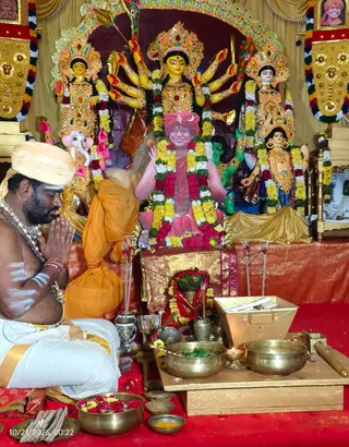 A photograph of a Navaratri puja at a shrine with a large Durga murti in the background, multi-armed and crowned, flanked by smaller painted deity figures and garlanded in marigolds and red flowers. In front, a kneeling priest in a white dhoti and cream turban with white tilak on his bare chest holds his palms together in namaste with eyes closed, wearing beaded necklaces. A painted or printed image of a figure in saffron-red robes and red headwear, decorated with a garland of yellow, white and red flowers, sits at the centre of the shrine. Brass bowls holding leaves, a brass container, flower bowls and lit oil lamps sit on a red cloth on the floor, and a saffron-clad figure sits to the left.