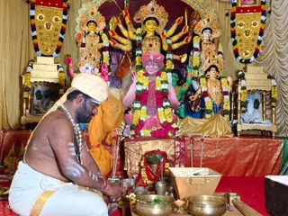 A photograph of a puja at a shrine decorated with a large multi-armed goddess murti flanked by two smaller deity figures, all garlanded with marigold and white flower strands. In the centre, a clay-like figure seated in a pink and red robe with a red turban and tilak, also garlanded. In the foreground a bare-chested priest in white dhoti and a white-and-cream head wrap, with white sacred ash markings on his body, kneels and performs ritual offerings beside brass bowls, a small oil lamp, incense sticks and flowers. A man in orange clothing stands behind him. Framed portraits decorated with garlands sit on side pedestals, and gold and red drapery frames the scene.