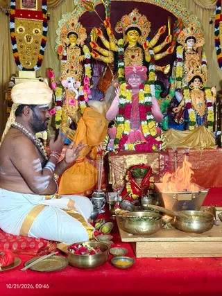 A photograph of a Navaratri puja at a shrine decorated with a large Durga murti with multiple arms, flanked by Ganesha and Lakshmi-style figures, all adorned with marigold and rose garlands. In front, a bare-chested priest in white dhoti with gold border and a cream turban sits on a red cloth, palms joined in prayer, with white marks on his arms and chest. Beside him a man in an orange garment is seated. A painted or printed figure in pink-red robes and a red turban with a crescent emblem sits on a throne at the centre, garlanded, its identity uncertain. Brass bowls of flowers, leaves, and offerings sit on the floor, with a small fire burning in a metal homa vessel to the right and a lit oil lamp nearby.