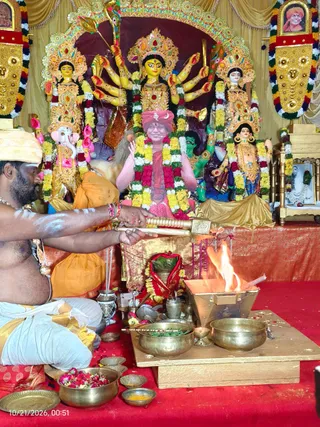 A photograph of a homa ritual in a decorated shrine room with a red floor cloth. A shirtless priest with white ash markings on his chest and arms, wearing a white dhoti with a gold border and an orange shawl, feeds offerings into a flame from a golden ladle over a brass fire pit. Brass bowls with leaves, flower petals and turmeric sit in front. Behind him stands a multi-armed deity murti with gold and red ornaments, flanked by a Ganesha figure and other images draped in garlands of yellow, white and red flowers. A framed portrait of a bearded man in an orange turban is at the upper right, and a seated figure in a red turban, pink and red garlanded clothing and a rudraksha mala sits in the center, appearing to be a statue or painted depiction rather than a living person. A timestamp reads 10/21/2026, 00:51.
