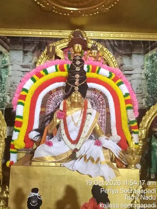 Photograph of a gold-coloured deity murti seated on a gold pedestal in a temple shrine, decorated with a striped garland arch of red, yellow, white and green flowers with pink fringe. The murti wears a white and gold-bordered dhoti and shawl, a red and white flower garland, gold necklaces and bangles, and a tall dark braided headdress adorned with gold ornaments. A brass kalasha-style lamp stands at the right, and a second small gold figure sits above the arch. Bright timestamped overlay reads Periya Seeragapadi, Tamil Nadu, kailasa seeragapadi.