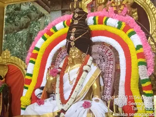 A golden statue of a seated figure, shown as a deity murti, is adorned for worship at a temple shrine. The figure wears a tall black crown-like headdress with gold ornaments, a white dhoti with a gold border, a gold necklace, and a long red and white flower garland. Its right hand is raised in a blessing gesture holding a small rudraksha mala. Behind it stands a large arched frame of concentric garlands in pink, yellow, red, green and white, with pink flowers at the top. A carved decorative backdrop and a painted forest scene appear in the background, and a timestamp overlay reads 19 Oct 2026 11:54:04 am along with Periya Seeragapadi, Tamil Nadu and kailasa seeragapadi.
