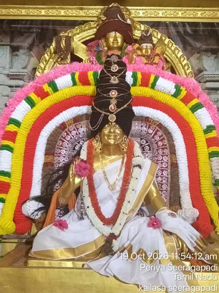 A photograph of a golden deity murti seated on a throne-like platform inside a decorated shrine, framed by a large arch of marigold and tuberose garlands in pink, yellow, red, green and white. The seated figure wears white and gold-bordered draped cloth, a long red and white flower garland, gold necklaces and earrings, and a tall black hair crown wrapped with gold ornaments and pendants, with a pink flower in the raised hand and a rudraksha-style mala hanging at the side. A smaller golden head with a red cloth wrap and a second small deity figure appear above. Printed white text on the lower right reads the date and time and location, and the scene is lit by warm light. Ritual decoration suggests a Navaratri festival darshan of the goddess Saraswati.