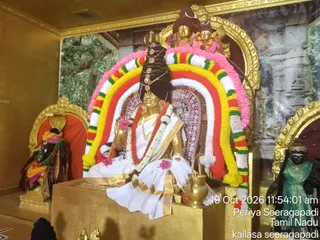 A seated golden deity murti, in a white and gold-bordered dhoti, is decorated with a red and white garland and a dark cloth crown. Behind it stands a large arch of garlands in pink, yellow, orange, red and white, framed by gold-edged niches. A small golden kalasha sits on the platform at its right side, and a dark figure in green and gold cloth stands at the right edge, with another dark figure and an orange-draped niche on the left. Printed timestamp and location captions appear in the lower right corner of the photograph.