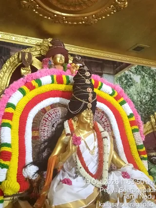 A photograph of a golden statue of a seated figure inside a temple-like shrine, decorated with a tall rainbow-coloured floral arch of red, yellow, white and green garlands edged in pink. The statue wears a black turban with gold ornaments and a pendant, a white dhoti, a red and white garland, a gold necklace and a pink flower in its hand. Behind it sits a smaller golden head figure in a maroon cloth, with rudraksha beads hanging on the wall. The background shows a gold-framed ceiling and a painted backdrop of trees. A timestamp overlay reads 19 Oct 2026 and Periya Seeragapadi, Tamil Nadu. No living people are visible.