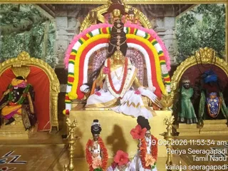 A photograph of a decorated shrine with a golden seated deity murti. A large arch of garlands in red, yellow, green and white with pink flowers frames the central figure. Flanking niches hold a red-curtained deity with a golden crown and a green-clad figure with a blue headdress. Two smaller decorated figures in red garlands and hibiscus flowers stand before the central platform, with brass oil lamps on either side. A painted forest backdrop completes the scene. The image carries a timestamp of 9 Oct 2026 and the location label Periya Seeragapadi, Tamil Nadu.