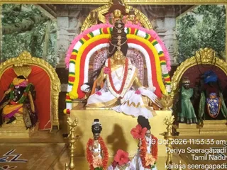 A goddess murti seated on a golden throne in a shrine decorated for Navaratri. She wears a white and gold sari, a red and white garland, and a tall ornate headdress with a dark braid behind her shoulders. A multicolored arched garland of red, yellow, green, white and pink frames her. Flanking alcoves hold smaller painted figures, one in green and one in blue. Brass oil lamps stand on the golden platform, and two smaller figures in red garlands and white dress sit in front. Painted forest and temple backdrop. Timestamp reads 19 Oct 2026 11:53:33 am, Periya Seeragapadi, Tamil Nadu, kailasa seeragapadi.