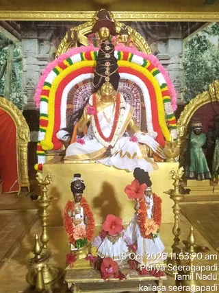 Photograph of a golden deity murti seated on a gilded pedestal inside a temple shrine, adorned with a multicoloured floral arch of pink, white, yellow, red and green garlands. The seated golden figure wears a white sari with gold borders, a red and white garland, and an elaborate dark braided headdress with jewelled ornaments. Flanking the pedestal are two smaller dark-faced murtis in white garments with orange-red flower garlands and hibiscus blossoms. Brass oil lamps stand on both sides, and a painted backdrop of temple architecture and greenery fills the background, with a green-clad figure visible on the right.