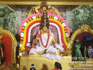 A seated golden deity murti of Saraswati stands at the centre of a shrine, dressed in a white sari with gold border and a red-and-white garland. A large arched backdrop of rainbow-coloured flower garland in pink, yellow, red, white and green frames the figure, and a tall dark braided hairstyle rises behind the head with a gold crown. Golden pots and a brass oil lamp sit at the base, with a golden peacock figure at lower left and painted forest and temple imagery on the walls. Smaller dark-skinned murtis dressed in green and blue stand in side niches at right. The timestamp reads 19 Oct 2026 11:52:51 am, with Kadathur Agraharam, Tamil Nadu and kailasa seeragapadi printed at lower right.