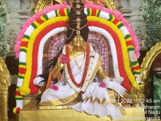 A golden deity murti of Goddess Saraswati seated on a gold pedestal, dressed in a white sari with a gold border and gold-trimmed orange under-cloth. She wears a gold necklace, bangles and a tall hair crown with gold ornaments. A thick garland of red, yellow, green, white and pink flowers forms an arch behind her, with pink blossoms placed at her hand and feet. A brass kalasha-style lamp stands at her right side and a green-clad figure is visible at the far right edge. Ornate gold-framed panels surround the scene. The photo is timestamped 19 Oct 2026 11:52:45 am at Kadathur Agraharam, Tamil Nadu, under the kailasa seeragapadi label, consistent with the Mahasaraswati darshan of the ninth day of Navarathri.