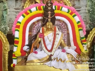 A golden deity murti of Goddess Saraswati seated on a gold pedestal, dressed in a white sari with gold border and an orange under-cloth. She wears layered gold necklaces, a red and white garland, and a tall dark hair crown adorned with round red stones. Behind her is a large arch of marigold-style garlands in red, yellow, green, white and pink, with pink flowers placed at her feet and lap. The shrine has ornate golden framing and a patterned backdrop. A date and location stamp reads 19 Oct 2026 11:52:42 am, Kadathur Agraharam, Tamil Nadu, kailasa seeragapadi.