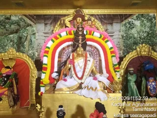 Photograph of a decorated deity murti seated on a gold pedestal inside a shrine, framed by an arch of garlands in pink, white, red, yellow and green. The central golden figure wears a white and gold sari with a red garland, a black and gold headdress with ornaments, and holds a gold kalasha-like vessel at its side. Flanking shrine niches on either side hold smaller dark-skinned figures in green and blue dress and red-orange garments. Painted tree-and-temple backdrop behind the shrine, with small offerings and a red hibiscus at the base. A timestamp reads 19 Oct 2026, 11:52:39 am, Kadathur Agraharam, Tamil Nadu, kailasa seeragapadi.