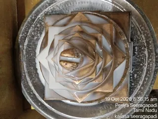A top-down photograph of a metallic sri yantra style geometric diagram, a set of nested triangles and squares with a central bindu in gold tones, placed on a large silver-toned tray or plate with a wet, glossy finish, possibly after abhishekam. No people are visible.