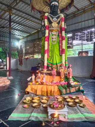 A photograph inside a covered open-sided hall showing a tall black stone deity murti standing on a raised platform, draped in a green silk sari with gold-bordered pallu, adorned with layered gold necklaces, a large pink and white flower garland, and a decorative headdress. Smaller figurines of deities and attendants stand on the orange and green cloth-covered platform, with flower petals scattered below. In front, a row of brass bowls filled with offerings sits on a green-bordered mat, alongside a silver tray with flowers and a lit oil lamp on a small white stand. No people are visible in the scene; a standing figure appears in the background at left.