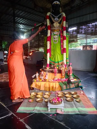 A photograph of a Deepavali neivedyam offering at a temple hall with a tin roof. A sadhika in an orange saffron sari, barefoot, stands at the left, holding a lit oil lamp in one hand while raising the other arm toward a large black deity murti. The tall black murti wears a green silk sari with a gold border, layered gold and silver necklaces, jewelled earrings, and long garlands of white and pink flowers. At its base on an orange and green silk-draped platform stand smaller painted deity figures adorned with garlands. In front, a row of brass bowls holds food offerings, and a central brass thali with pink flowers and lamps sits on a small white stool atop a green and pink patterned cloth. Scattered petals lie on the dark stone floor.