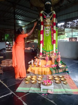 A woman in an orange sari stands at the left of a large black deity murti draped in a green sari, garlanded with white and pink flower strings and wearing a gold necklace and crown-like headdress. She reaches toward the image with one hand while holding an offering or lamp in the other. Smaller murtis and flowers sit on an orange and green cloth-draped platform, with rows of brass bowls filled with offerings and a brass plate of petals on a small white stool in front. The setting is a covered open hall with a metal roof and dark tiled floor.