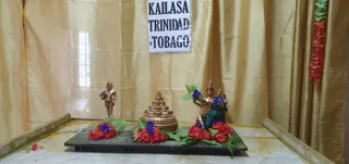 A photograph of a temple shrine set against gold curtains. A paper sign reading "KAILASA TRINIDAD & TOBAGO" hangs at the top centre. On a dark wooden platform sit three bronze-coloured murtis: a standing figure at left holding a blue flower, a small bronze shrine-like structure with stepped tiers in the centre, and a seated deity murti at right wearing a green cloth and holding a blue flower, with a smaller figure beside it. Red and blue flower garlands and green leaves decorate the base. No people are visible.