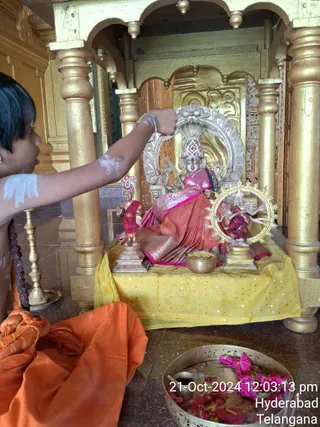 A young boy with a bare upper body, orange cloth wrapped around his waist and white sacred markings on his arm and shoulder, reaches up with his hand to touch or offer to a seated deity murti in a gold-pillared shrine. The murti wears a pink and red silk sari with a crown, seated on a yellow sequined cloth-covered platform. Smaller figures stand beside it, one in red holding a small object and one in a circular gold halo. A brass bowl of offering sits in front, and a brass tray holds pink flowers and rose petals in the lower right. Overlaid timestamp reads 21-Oct-2024 12:03:13 pm, Hyderabad, Telangana.
