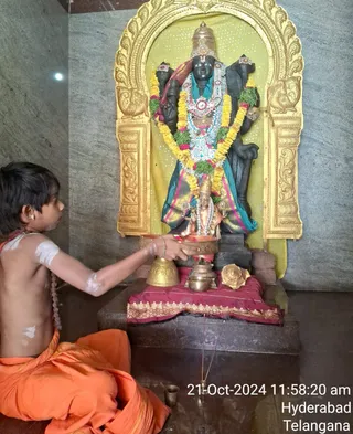 A young boy with bare upper body and orange dhoti sits cross-legged on the left, holding a brass bowl toward a black stone deity murti dressed in a teal and gold silk garment, adorned with yellow and green garlands, a jewelled necklace and a gold crown. In front stand a brass bell, a brass kalasha-style pot, a conch shell and a small brass vessel on a maroon cloth with gold border, with incense sticks burning. The murti sits in a gold-framed arched niche against grey stone walls, with a small brass lamp on the floor. Scene suggests a puja or abhishekam in progress, with white marks painted on the boy's shoulders and arms. A timestamp reads 21-Oct-2024 11:58:20 am, Hyderabad, Telangana.